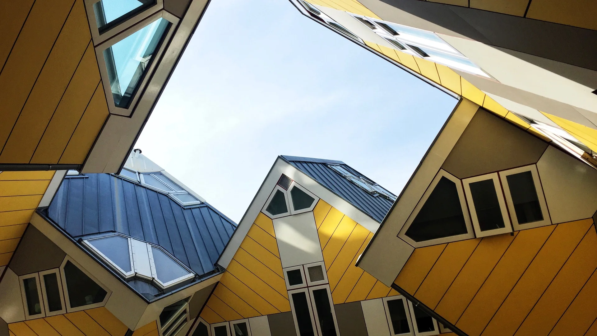 Yellow cube shaped houses tilted onto their corners, photographed from below against a pale blue sky, with gray slate roofs and white framed windows set into the slanted faces.