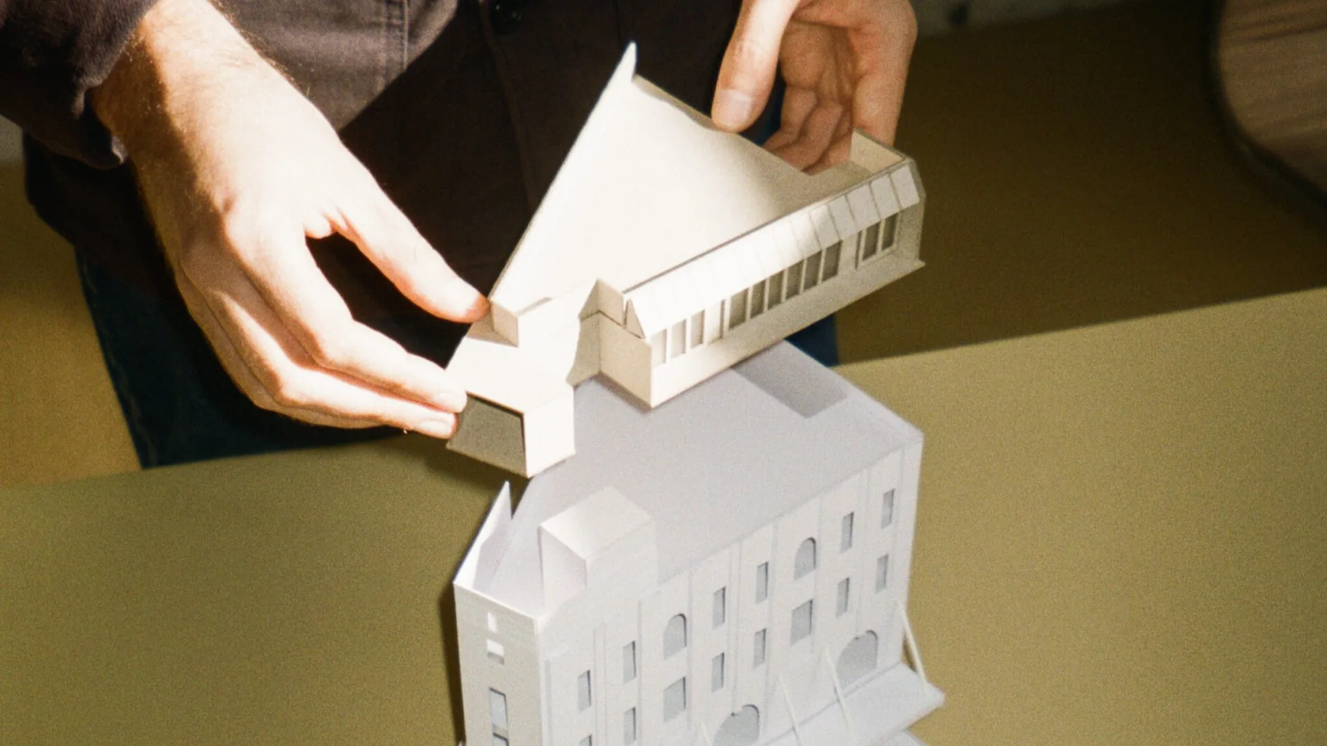 Two hands lifting a white paper architectural model of a roof section above a larger white paper model of a building with arched windows, on an olive green table.