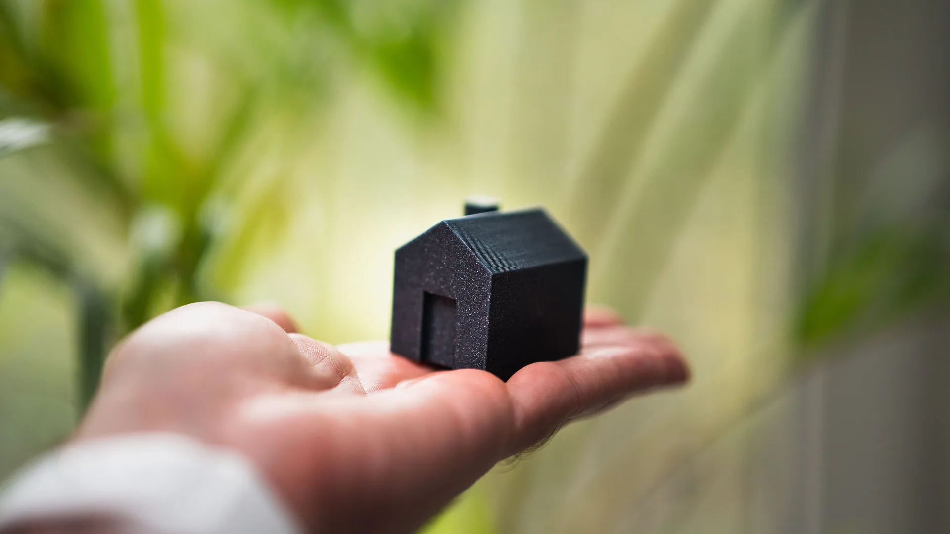 An open hand holds a small dark model house against a blurred green background.