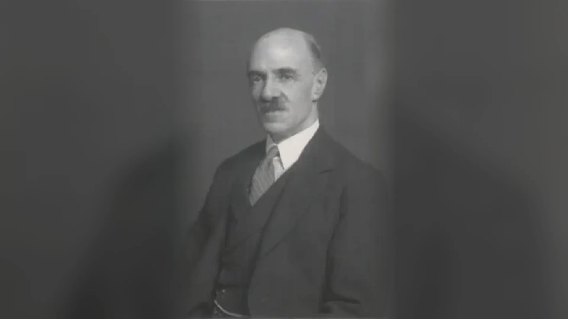 A black and white studio portrait of an older bald man with a moustache, seen from the chest up in a dark three piece suit, white shirt and striped tie, against a plain grey background.