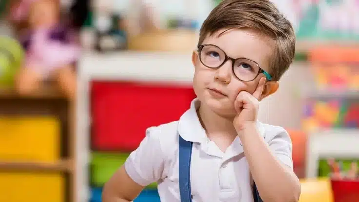 A young boy wearing round glasses and a white polo shirt rests his chin on one hand in a brightly colored classroom.