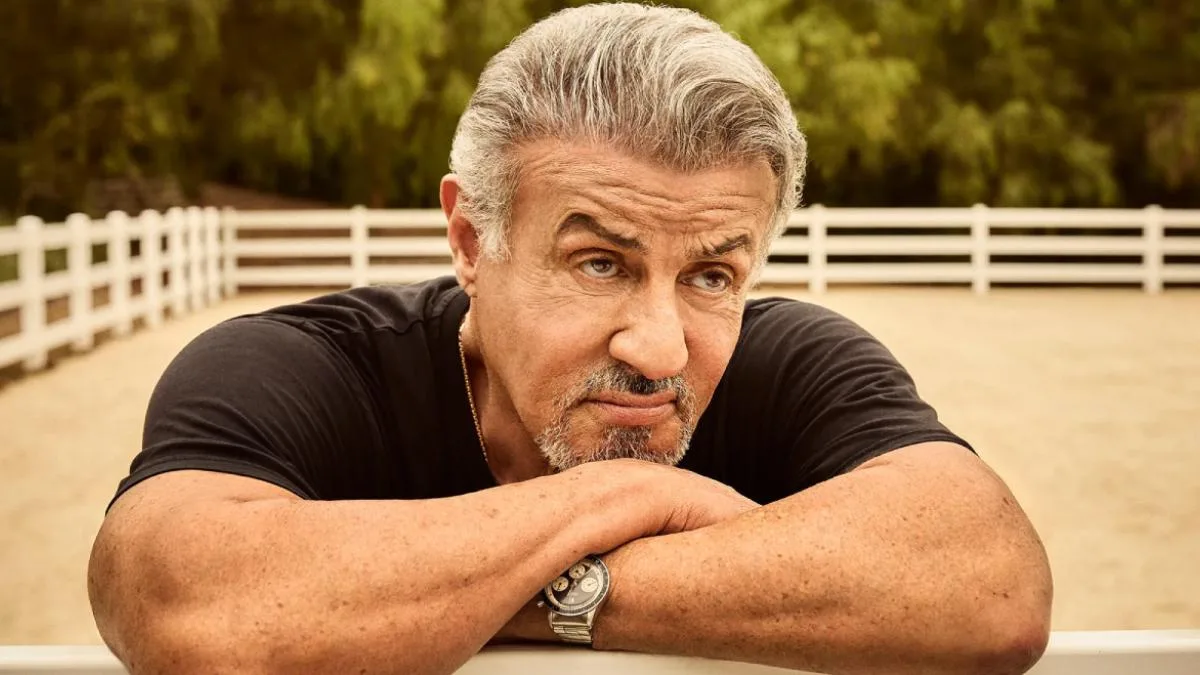 Sylvester Stallone in a black t-shirt, gray hair swept back, leaning on a white fence rail with his arms folded.