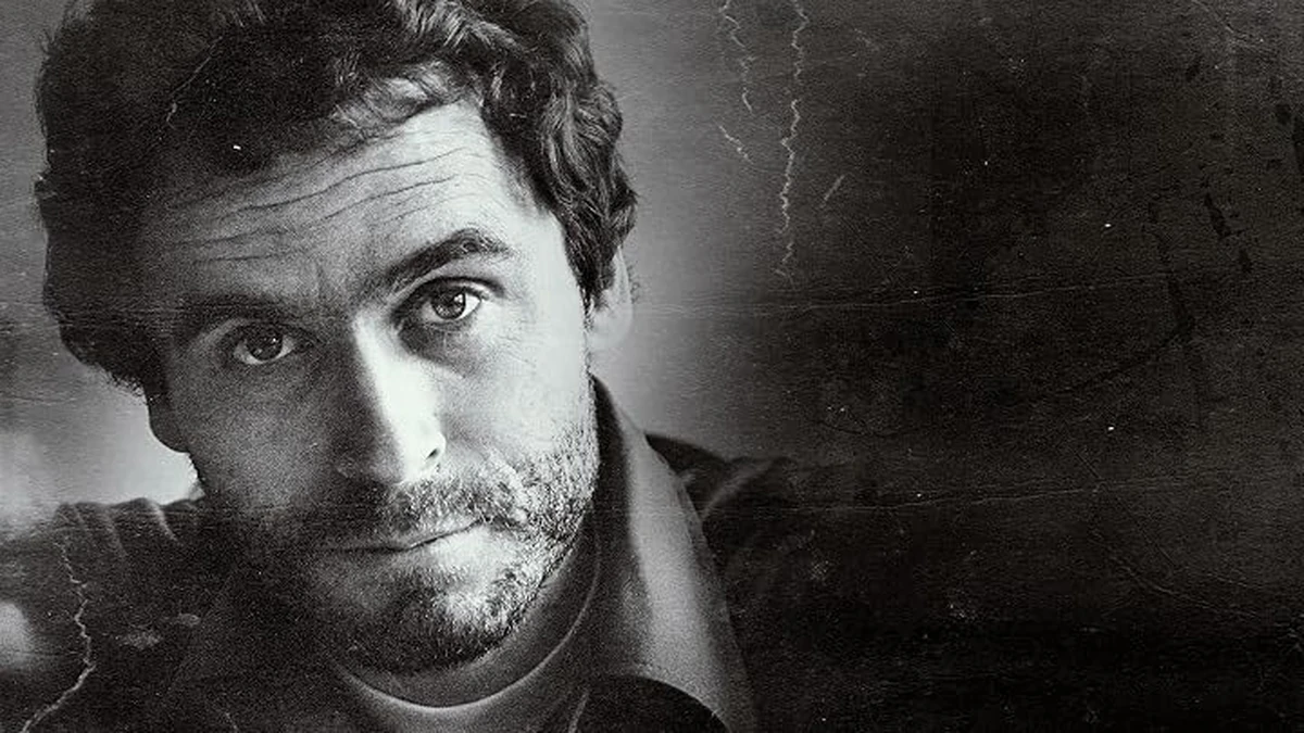 Black and white close-up photograph of a man with dark curly hair and stubble, his head tilted slightly as he looks toward the camera, wearing an open-collared shirt against a scratched, dark gray background.