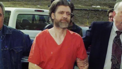 Ted Kaczynski in a red jail jumpsuit escorted by officers past parked vehicles outdoors.