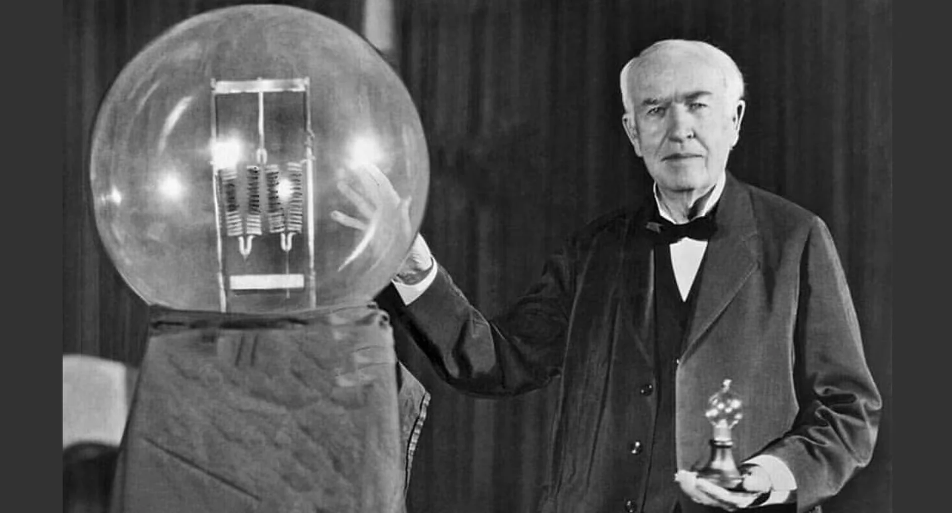 Black and white photograph of an older man with white hair, in a dark suit and bow tie, resting one hand on a large clear glass globe that holds a coiled filament assembly while he holds a small light bulb in the other hand, in front of a dark curtain.