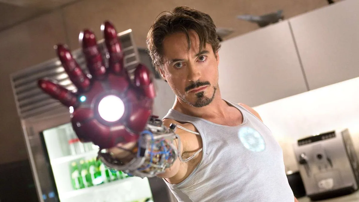A man with dark hair and a goatee, wearing a gray sleeveless shirt with a small round glowing light on the chest, holds out a red armored glove with a glowing white palm and exposed wiring toward the camera, in a kitchen with a refrigerator behind him.