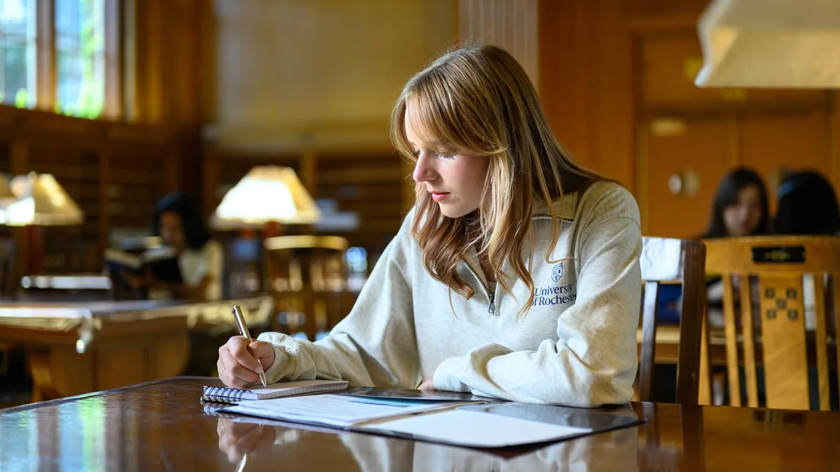 A young woman writes in a notebook at a wooden table in a library reading room lit by lamps.
