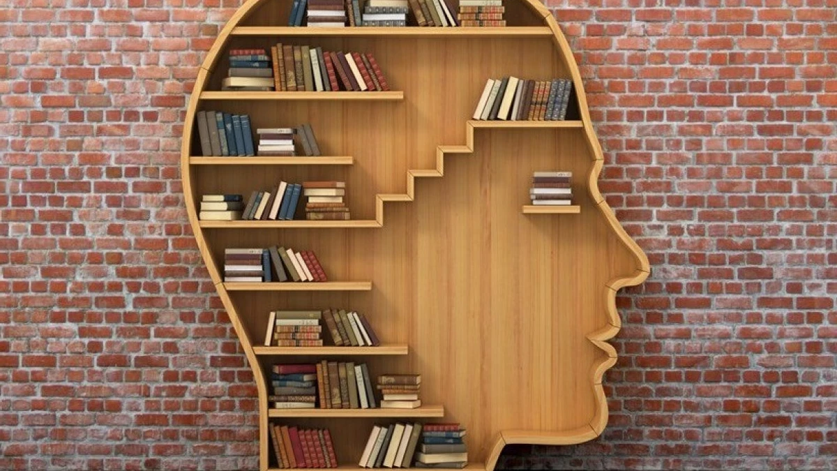 A wall mounted wooden bookshelf cut in the shape of a human head in profile, its shelves filled with rows of hardback books, fixed to a red brick wall.