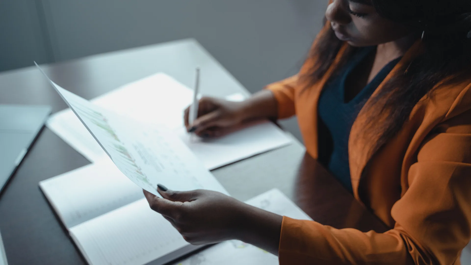 A woman in an orange blazer holds a printed sheet in one hand and a pen in the other above an open notebook.