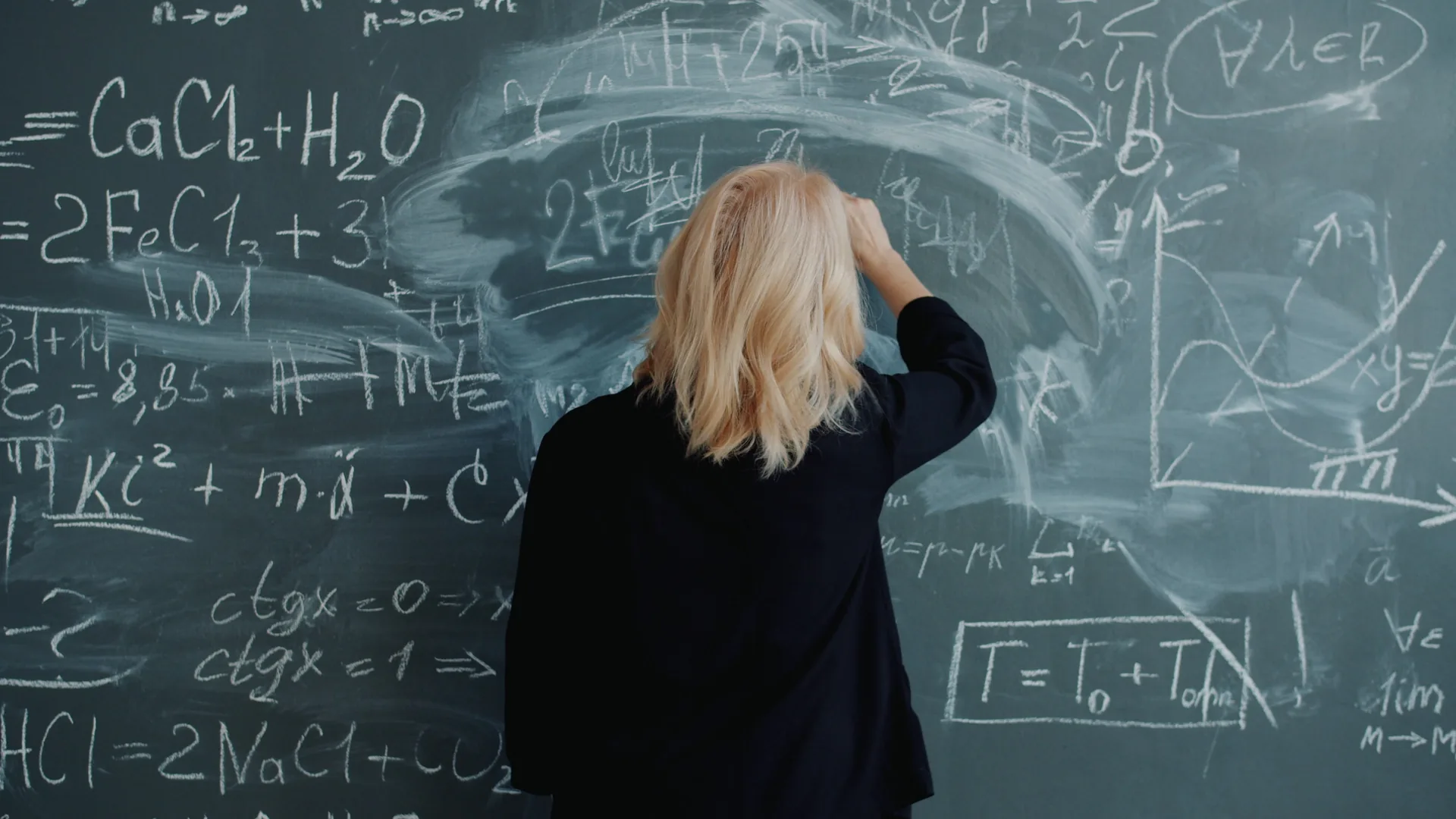 A woman with shoulder-length blond hair, seen from behind in a dark top, raises her right hand to a green chalkboard covered in chalk equations, chemical formulas and graphs, with a wide smeared patch where part of the board was wiped.