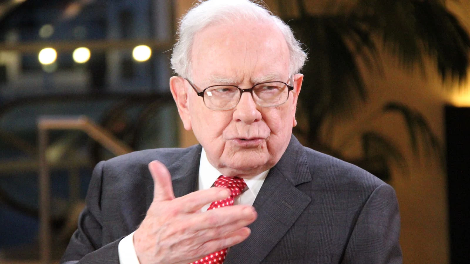 Elderly man with white hair and glasses, in a dark suit, white shirt and red polka dot tie, gesturing with one hand while he speaks, in front of a blurred indoor background with warm lights and a dark palm leaf.