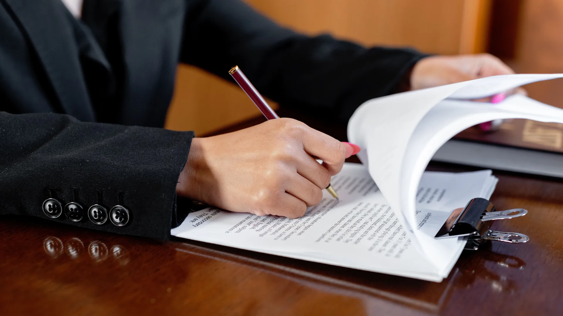A person in a dark suit writes with a pen on the pages of a document at a polished wooden desk.