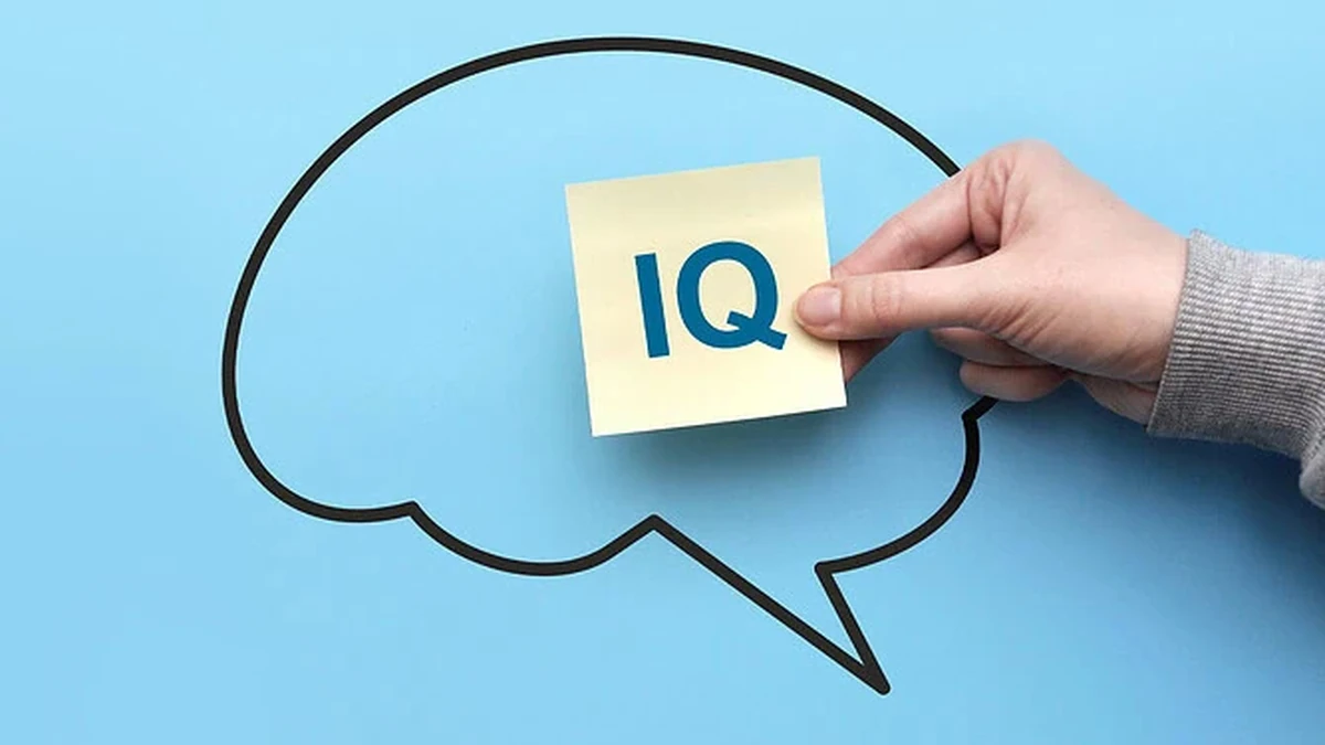 A hand holds a pale yellow sticky note marked IQ in blue letters in front of a thick black outline of a brain drawn as a speech bubble, on a light blue background.
