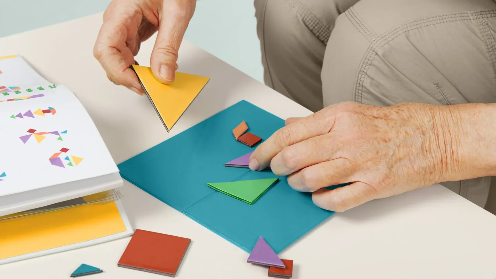An older adult's hands arranging yellow, green, purple and red card triangles and squares on a teal board beside an open stimulus booklet.