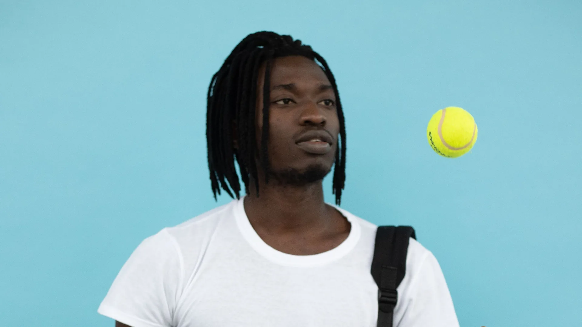 A young man with a backpack strap on his shoulder watches a tennis ball in the air against a blue wall.